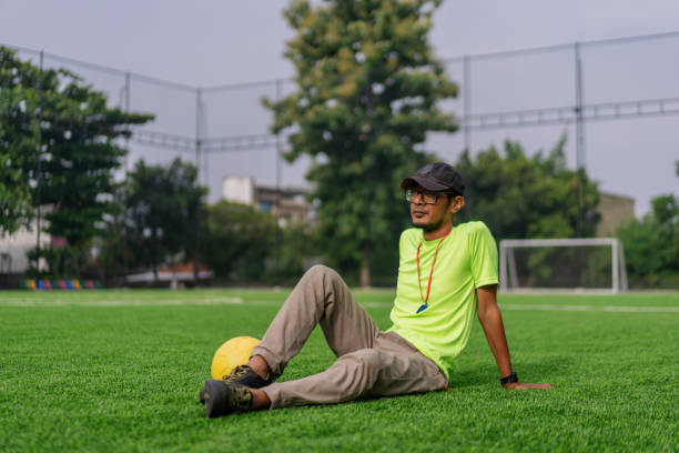 A male soccer coach wearing a neon yellow sports shirt, khaki pants, and a black cap sits on the artificial turf field with a relaxed posture. A whistle hangs around his neck, and a yellow soccer ball rests near his foot.