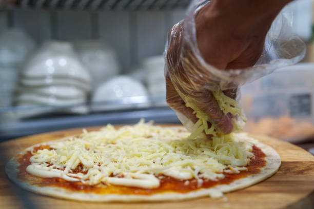 A person wearing gloves preparing a homemade pizza by sprinkling shredded cheese on a thin crust with tomato sauce and mayonnaise in a kitchen setting.