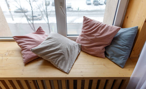 Four pillows are on a wooden bench by a window. The pillows are pink, white, and blue. The bench is next to a radiator