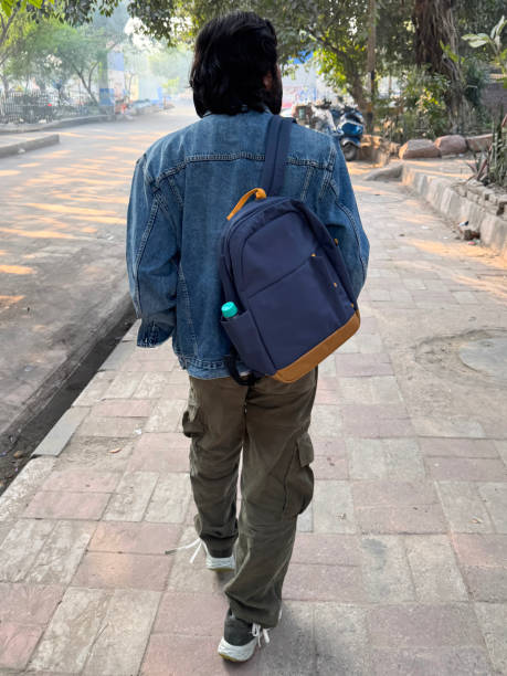 istock Image of man walking on pavement with backpack, blue denim jacket and carrying water bottle / Man walking on urban street 2202209769