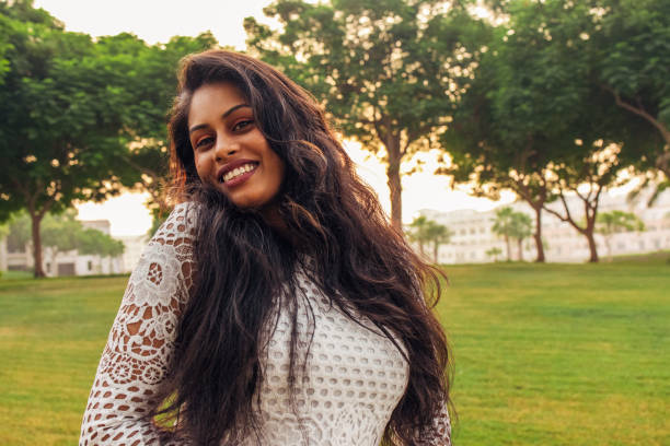 A young woman poses gracefully in a green park, her arms elegantly framing her face as she smiles warmly. Dressed in a delicate white lace outfit, she radiates charm and confidence against the serene backdrop of lush trees and open space. The soft natural light enhances her glowing complexion, adding a touch of magic to this lighthearted and artistic moment.