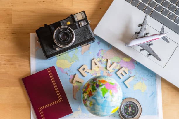 World map with globe, compass, passport, camera and laptop displaying a miniature airplane on top of a wooden table suggest planning and booking the next summer vacation trip