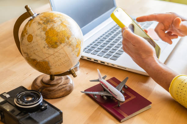 Travel agent using a smartphone and laptop for booking trips, surrounded by a globe, passport, toy airplane, and vintage camera on the desk, embodies modern travel planning