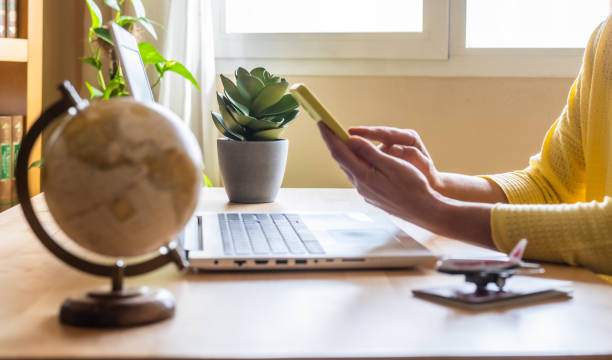 istock Travel agent browsing on a mobile phone and working on a laptop while booking a vacation, surrounded by a globe, airplane model, and passport on the desk 2202212012