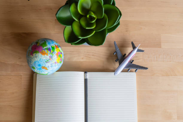 Open notebook with a pencil lies on a wooden table next to a small globe, a model airplane, and a potted plant, suggesting travel planning and wanderlust