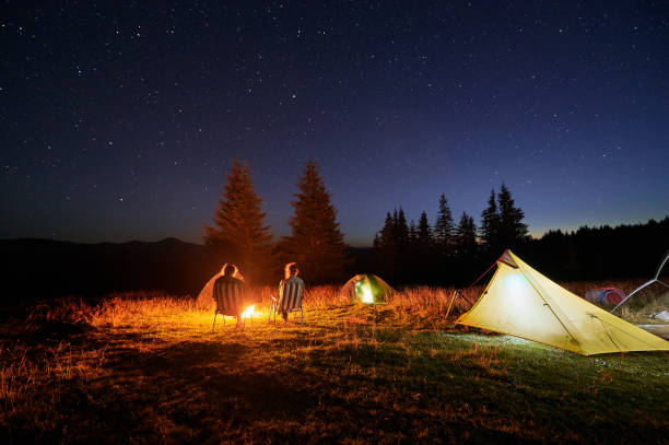 istock Two people campers sit by campfire under starry sky, surrounded by illuminated tents. 2202219604