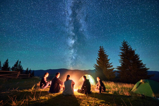 istock Group of hikers resting around campfire, surrounded by illuminated tents under stunning starry sky. 2202219608