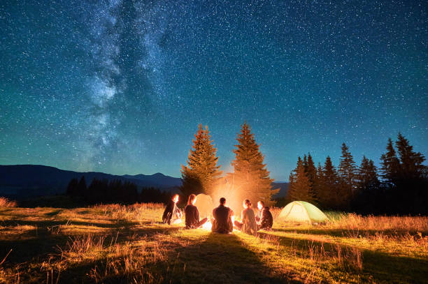 istock Group of hikers resting around campfire, surrounded by illuminated tents under stunning starry sky. 2202219609