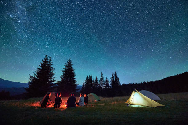 istock Group of hikers resting around campfire, surrounded by illuminated tents under stunning starry sky. 2202219616