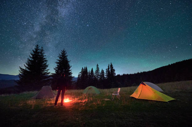 istock Person hiker having a rest by campfire, surrounded by glowing tents under magnificent starry sky. 2202219673