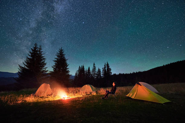 istock Person hiker having a rest by campfire, surrounded by glowing tents under magnificent starry sky. 2202219681