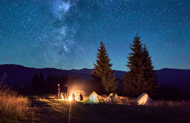 istock Group of hikers resting around campfire, surrounded by illuminated tents under stunning starry sky. 2202219683
