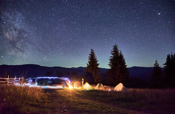 istock Group of hikers resting around campfire, surrounded by illuminated tents under stunning starry sky. 2202219686