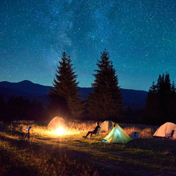 istock Person hiker having a rest by campfire, surrounded by glowing tents under magnificent starry sky. 2202219691