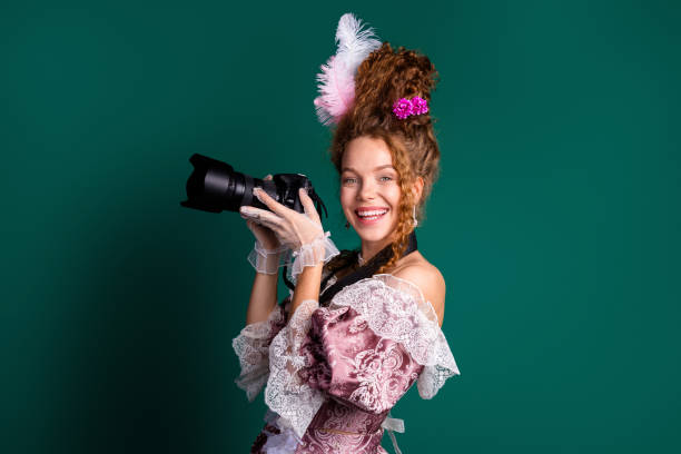 istock Young woman in baroque costume holding camera against green backdrop, blending elements from eras past and modern styles 2202223976