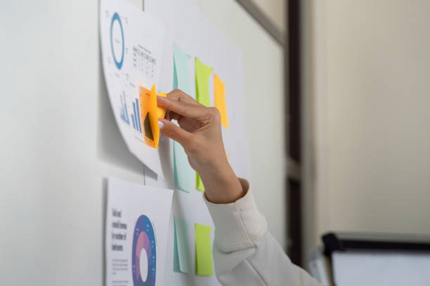 istock Hand placing sticky notes on a wall filled with financial charts and documents, promoting organization. 2202238887
