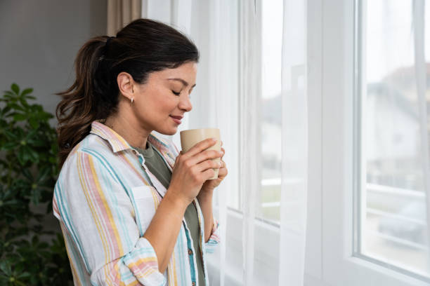 istock Beautiful young woman drinking a cup of coffee while looking forward standing next to the window at home. Morning routine and waking up with hot beverage. Relaxation and happiness. 2202241726