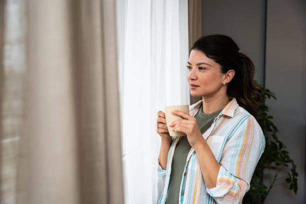 istock Beautiful young woman drinking a cup of coffee while looking forward standing next to the window at home. Morning routine and waking up with hot beverage. Relaxation and happiness. 2202241742