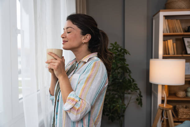 istock Beautiful young woman drinking a cup of coffee while looking forward standing next to the window at home. Morning routine and waking up with hot beverage. Relaxation and happiness. 2202241743