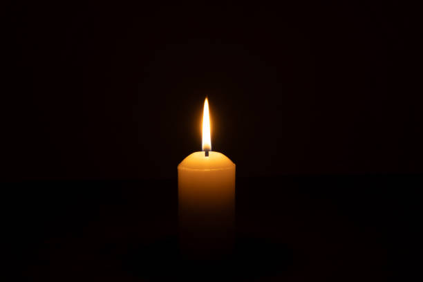 istock A single burning candle flame or light glowing on a big white candle isolated on black or dark background on table in church for Christmas, funeral or memorial service 2202242726