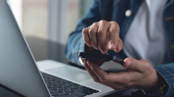 istock Closeup, business woman sitting at table using mobile smrt phone during working on laptop computer at home office. Young asian woman freelancer  surfing the internet, online working from home 2202245033