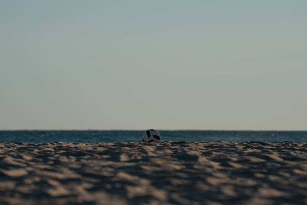 White soccer ball resting on the sandy beach, with the serene sea and horizon in the background, creating a peaceful summer scene