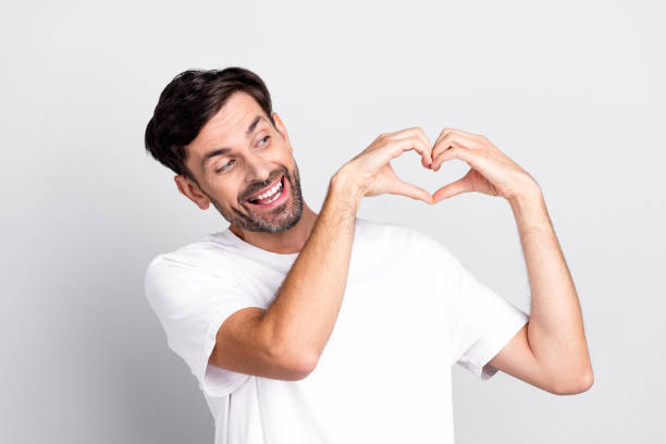 istock Smiling man making a heart hand gesture while wearing a casual white shirt on a gray background conveying happiness and a loving mood 2202250376