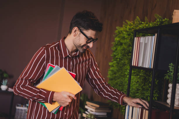 istock Young man in striped shirt selecting a book from the shelf while holding colorful notebooks, embodying a student lifestyle 2202252272