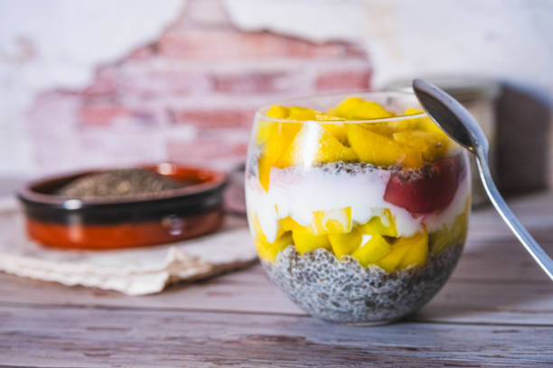 A glass of fruit salad and chia pudding with a spoon in it. The fruit salad is made with bananas, mangoes, and strawberries