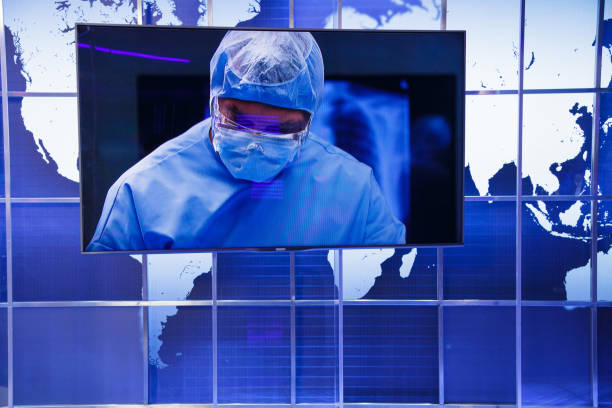 A modern TV news studio background with a blue-lit panel wall featuring an earth map, symbolizing global news and communication. A large display screen on the wall shows a medical professional wearing a protective suit and safety glasses, possibly a surgeon, bio lab researcher, or infectious disease expert. The futuristic blue tones and high-tech design create a serious and informative atmosphere, representing science, healthcare, and global media coverage.