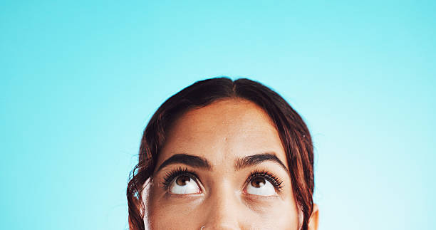 Closeup, eyes and woman in studio, thinking and problem solving on blue background. Person, looking up and model with ideas, thoughtful and solution with decision, option and choice with mockup space