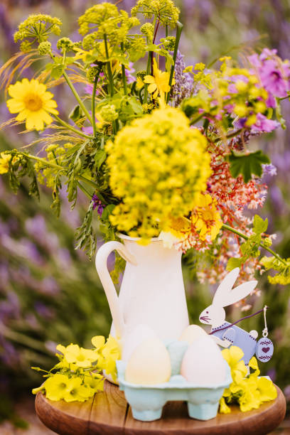 Beautifully arranged Easter still life featuring a retro-style Easter bunny figurine beside a white porcelain milk jug repurposed as a vase holding a fresh wildflower bouquet. Easter eggs and delicate clover blossoms add to the festive charm. Color editing. Part of a series. Objects are decoration mass product.