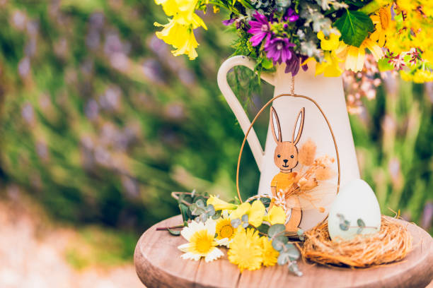 Beautifully arranged Easter still life with a gold-colored hanger displaying a wooden Easter bunny clutching dried flowers gently swaying in front of a white porcelain milk jug filled with meadow blooms. A small nest with an Easter egg sits beside it, completing the delicate composition. Color editing. Part of a series.Objects are decoration mass product.