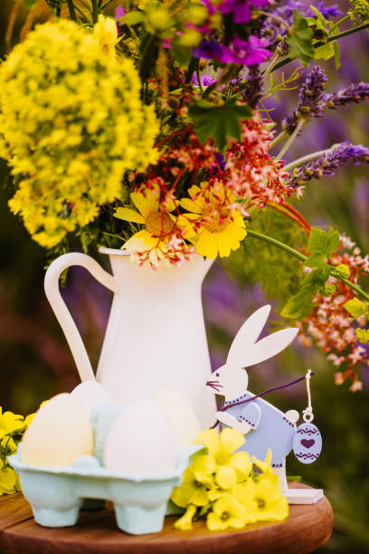 Lovingly decorated Easter still life featuring a vintage-style Easter bunny figurine next to a white porcelain milk jug used as a vase filled with a wildflower bouquet. Delicate Easter eggs and clover blossoms complete the charming seasonal arrangement. Color editing. Part of a series. Objects are decoration mass product.