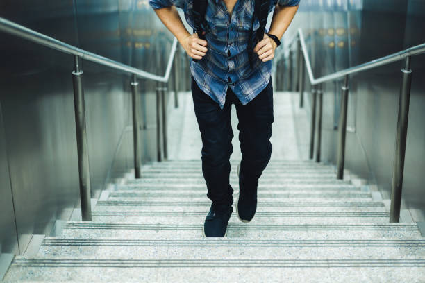 A young man wearing a plaid shirt and backpack walks up a stairway in an urban environment with metal railings and walls.