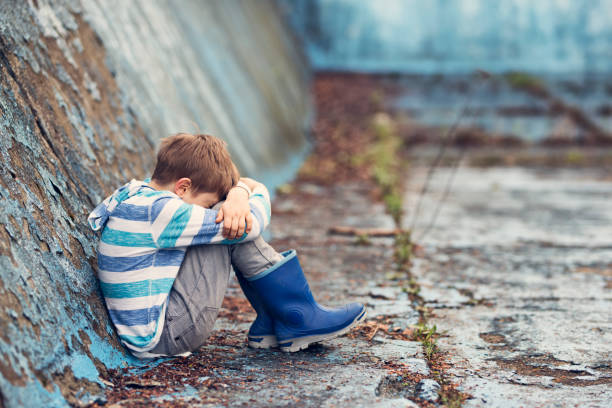 Young boy sitting alone at the bottom of an abandoned pool. The boy is wearing a striped hoodie, gray pants, and blue rain boots. The pool's surface is cracked and weathered, with patches of blue paint and overgrown weeds, indicating neglect and abandonment. The depressed boy is sitting with knees drawn up to the chest and arms wrapped around the knees. \nShot with Nikon D810