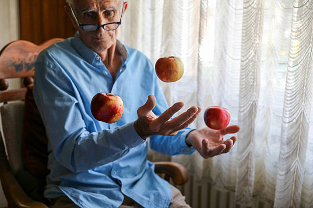 A senior man skillfully juggling several apples against a warm brown background, with a joyful expression on his face.