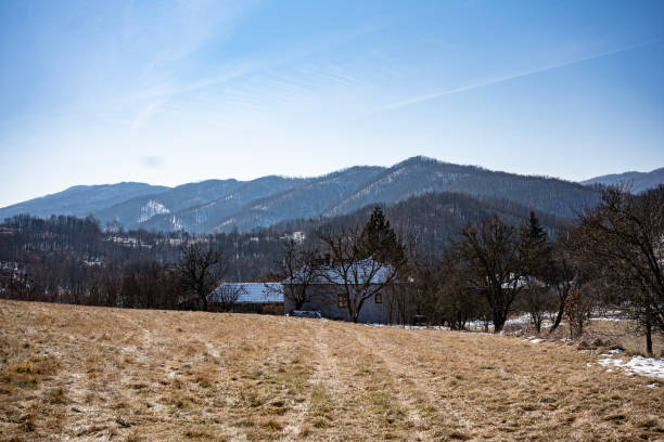 Tranquil rural winter scene featuring a lonely cottage surrounded by bare trees against a backdrop of snow-dusted hills and a bright blue sky.