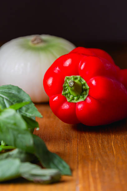 A white onion, a red bell pepper with water droplets, and fresh basil leaves, placed on a wooden table with a dark background.