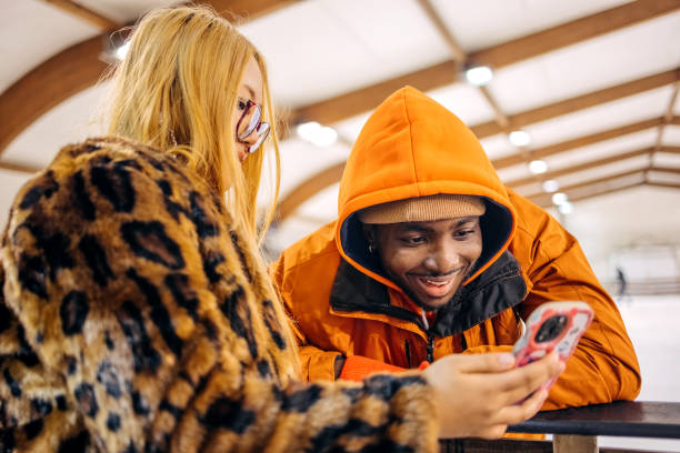 Young couple having fun ice skating indoor during winter and taking a break looking at mobile phone