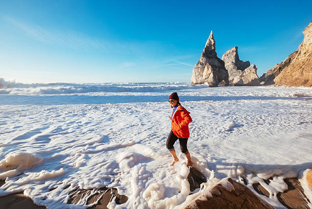 Lonely woman dressed red jacket walking by Beautiful abandoned Atlantic ocean Ursa beach and enjoying coastal surf with foamy waves crashing on rocky cliffs on Winter February coast in Portugal.