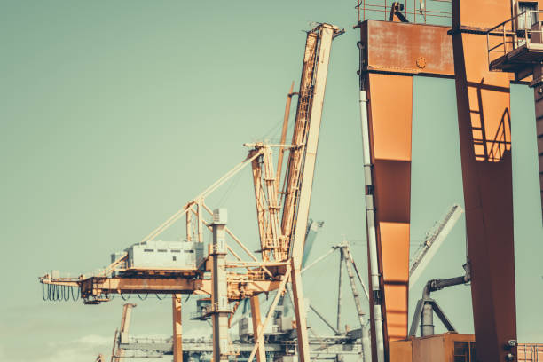 Industrial port scene with large orange gantry cranes and container handling equipment under a clear sky. Steel structures, cables, and shadows create a dynamic composition; retro colours, telephoto
