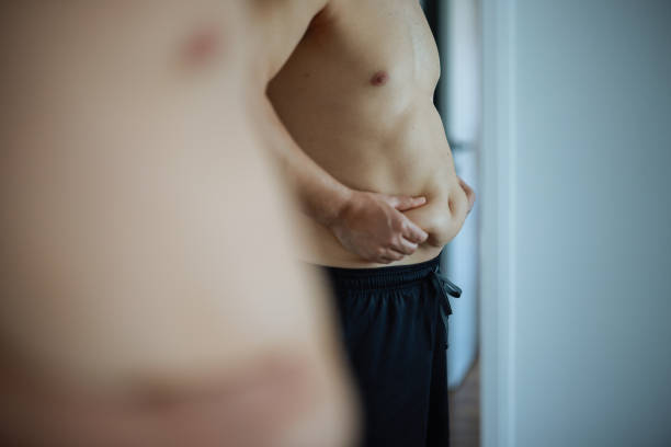 Upset man has extra weight looking at mirror touching big abdomen in bedroom at home. Bearded guy dissatisfied body shape. Obesity, overweight, healthy problems, weight control, bad unhealthy habits.