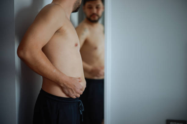 istock Overweight man checking his fat body reflection in mirror 2202339154