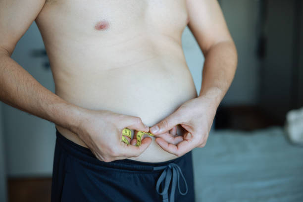 Upset man has extra weight looking at mirror touching big abdomen in bedroom at home. Bearded guy dissatisfied body shape. Obesity, overweight, healthy problems, weight control, bad unhealthy habits.