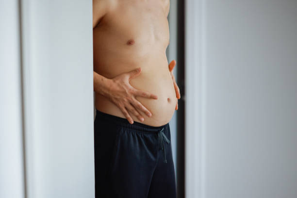 Upset man has extra weight looking at mirror touching big abdomen in bedroom at home. Bearded guy dissatisfied body shape. Obesity, overweight, healthy problems, weight control, bad unhealthy habits.