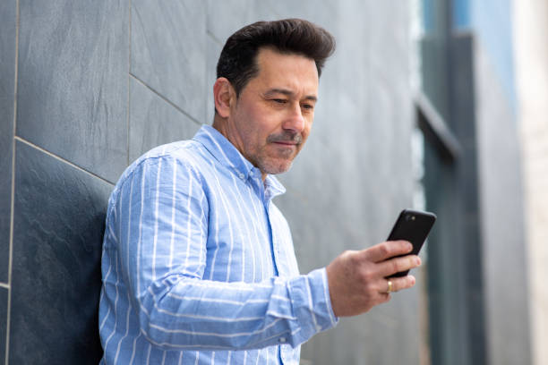 istock Older man leaning against wall looking at mobile phone 2202381660