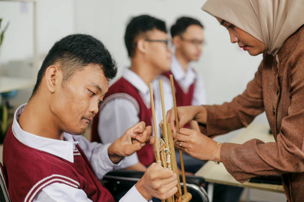 Young Female Teacher Helping Student With Physical Disability Making Angklung, Traditional Music Instrument From Bamboo.