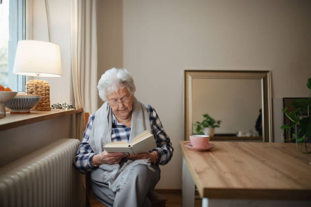 istock Lovely elderly lady reading a book, sitting at the kitchen table and sipping tea. 2202430527