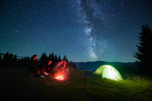 istock Group of friends hikers rests around campfire next to tent and solar panel under star-filled sky. 2202433214